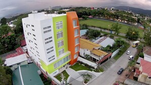 A multi-story building with a white and orange facade, surrounded by greenery and a sports field. - Hotel Latitud 15 (San Pedro Sula)