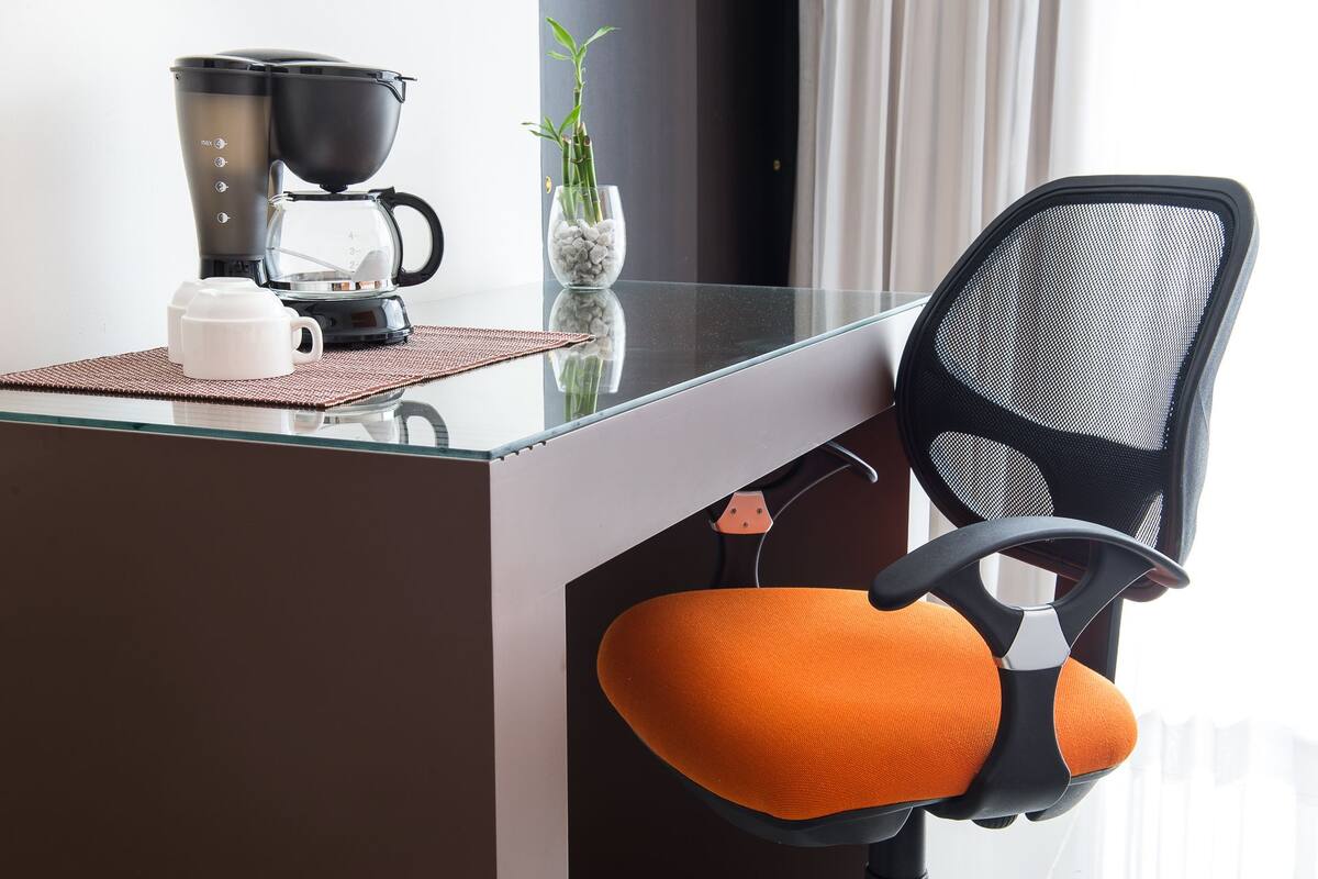 A modern office chair with an orange cushion, a glass coffee table, a black coffee maker, and a small potted plant.