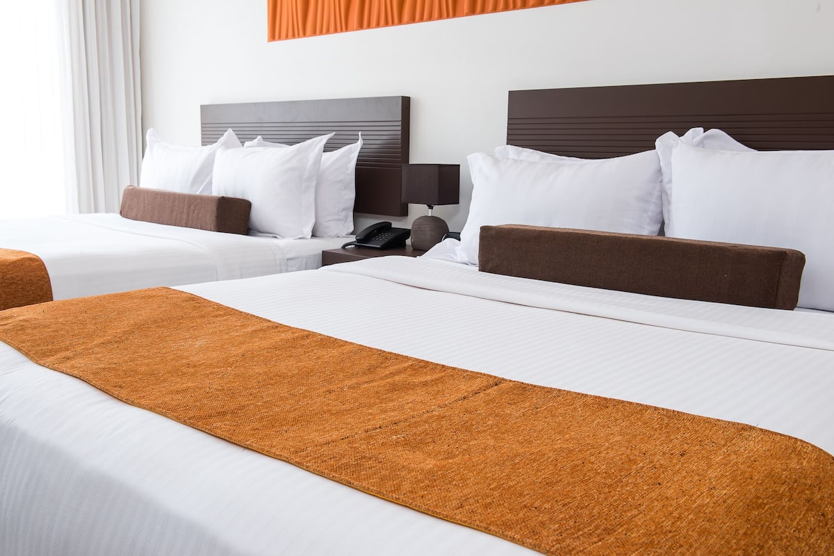 A hotel room with two beds, each with white bedding and brown headboards. There is a brown throw blanket on the bed in the foreground.