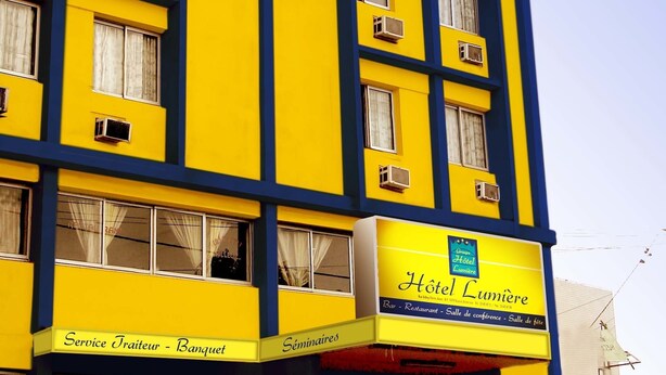 A multi-story hotel with a yellow facade and blue accents. It features air conditioning units, a sign for 'Hotel Lumière' with services like a bar, restaurant, and conference room. - Hôtel Lumière D'Akwa (Douala)