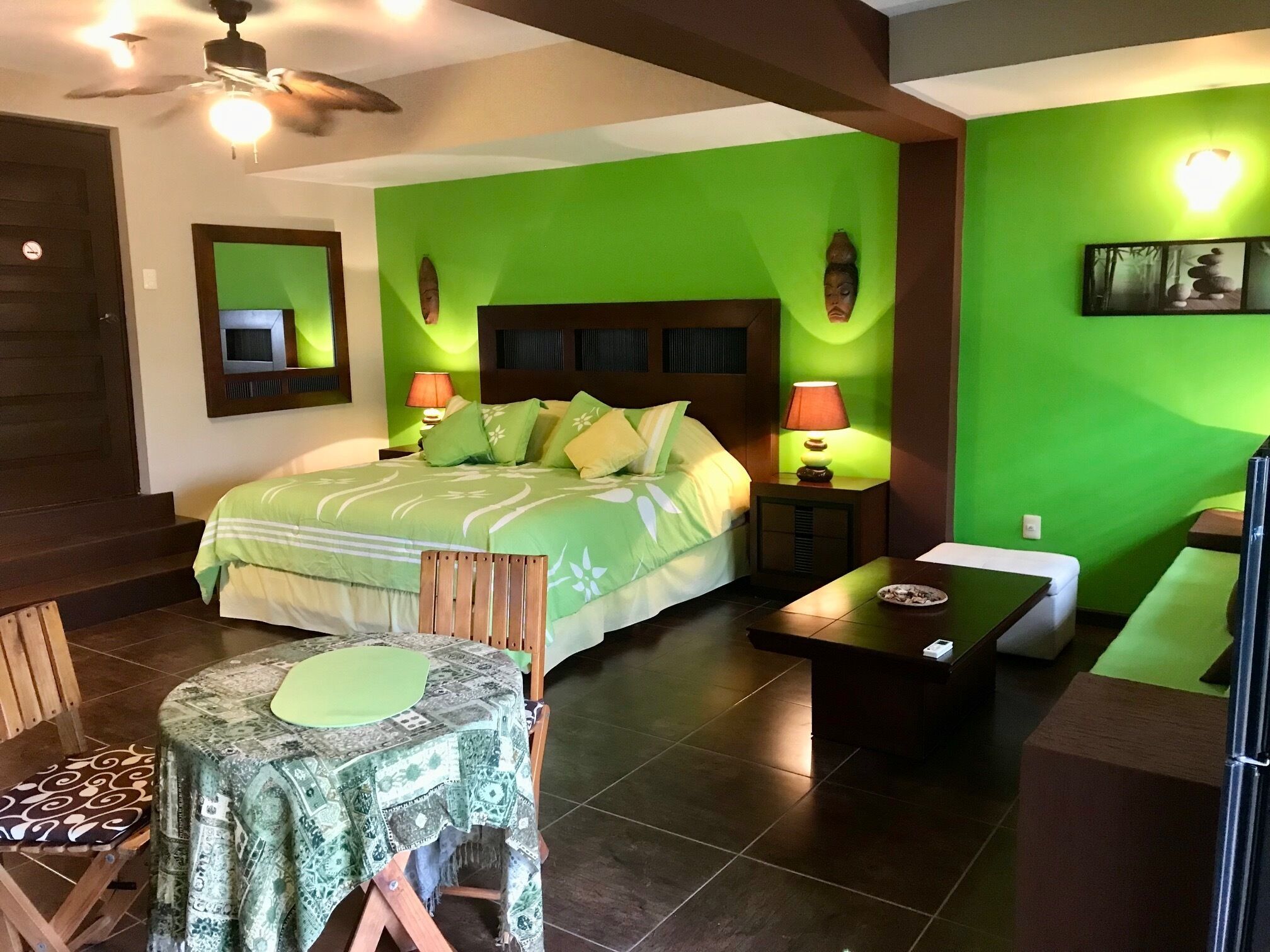 A bedroom with a green wall, a bed with green bedding, two bedside lamps, a wooden nightstand, a small table with a green tablecloth, and a chair with a patterned cushion.