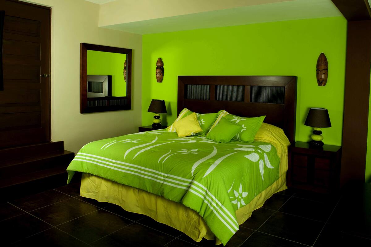 Een slaapkamer met een groene muur, een houten hoofdbord, een bed met groen en geel beddengoed, twee nachtkastjes met lampen, en een spiegel aan de wand.