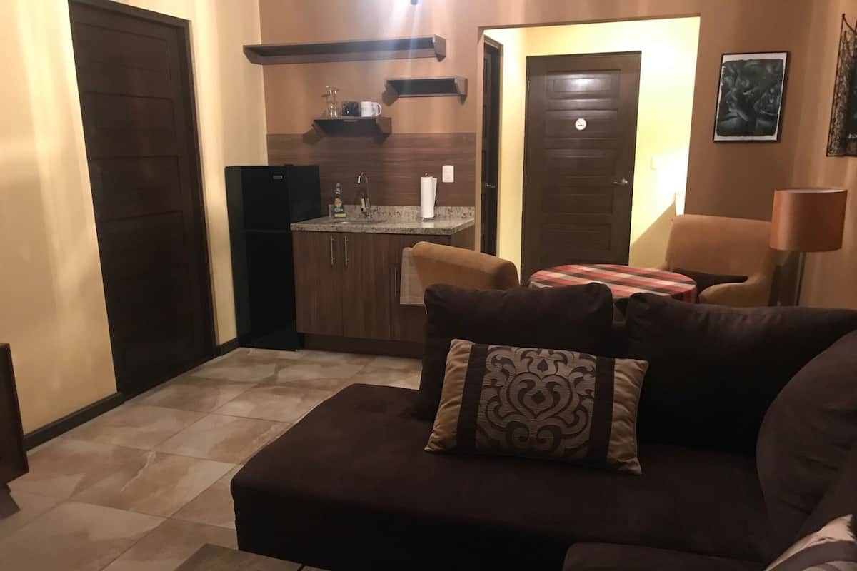 Een woonkamer met een donkerbruine bank, een salontafel en een sierkussen met een patroon. In de achtergrond is een keuken te zien met een spoelbak en kastjes.