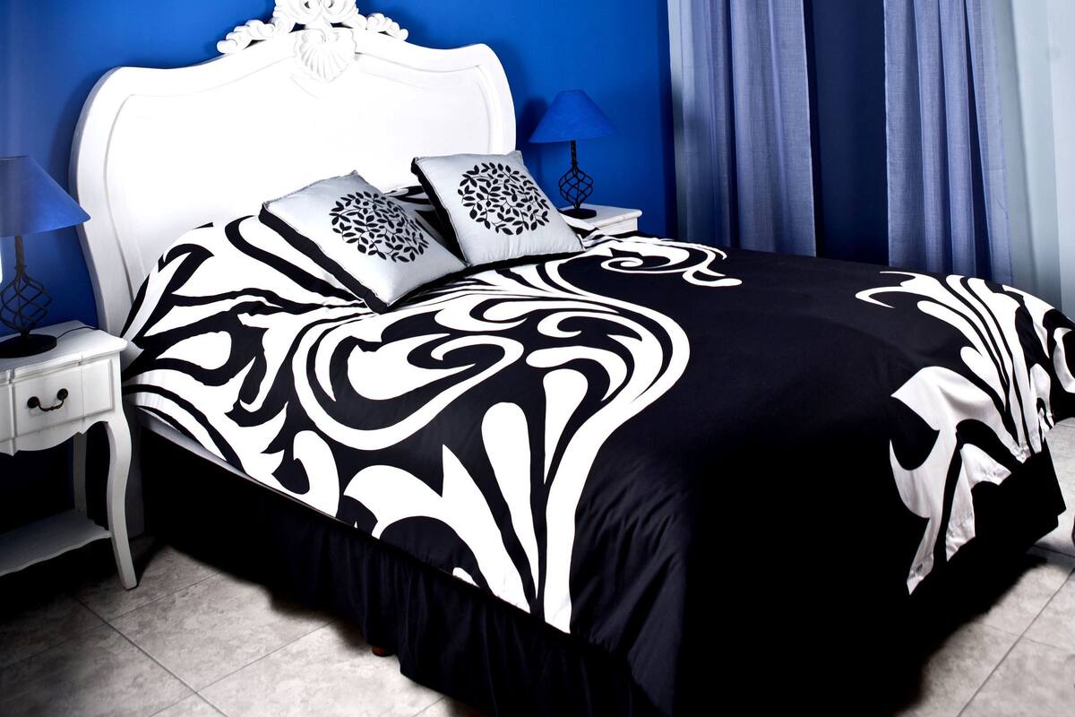 Een slaapkamer met een wit hoofdeinde, een zwart-wit beddegoed, een kroonluchter en blauwe gordijnen.