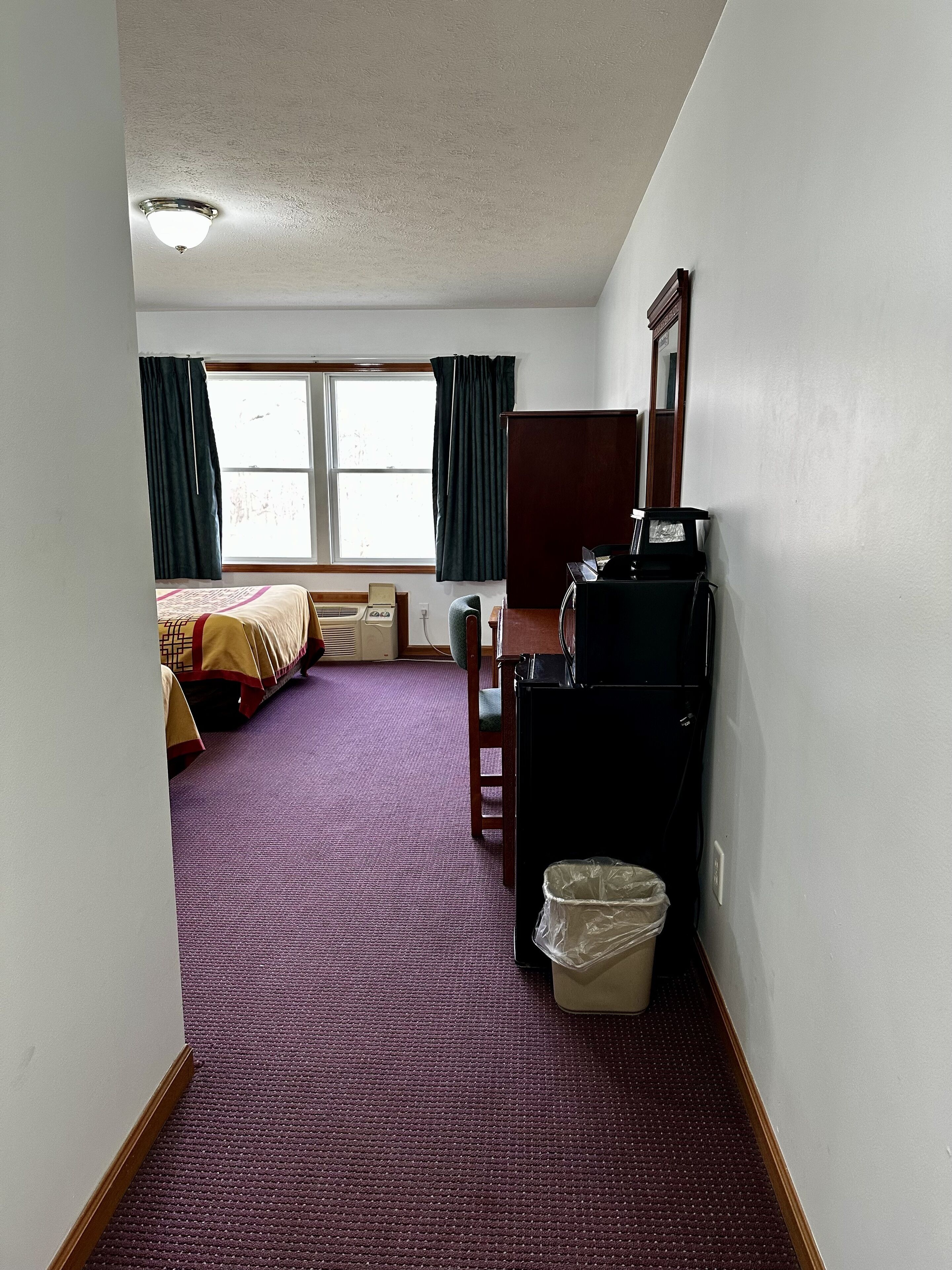 Habitación de hotel con alfombra púrpura, cama con colcha estampada, escritorio con computadora y ventana con cortinas.