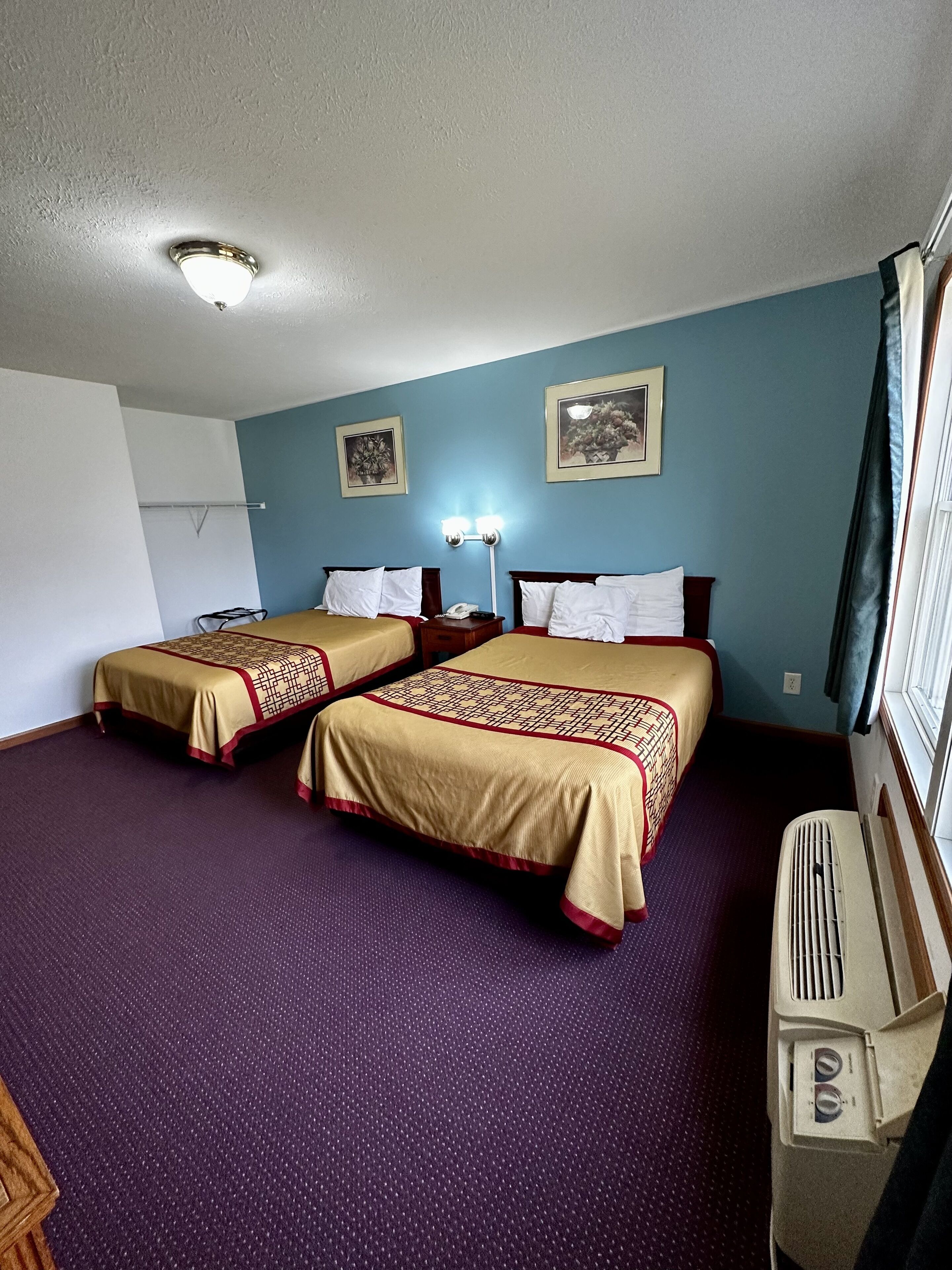 A hotel room with two beds, a ceiling light, a wall-mounted phone, and a window with curtains.