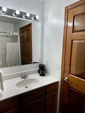A bathroom with a white sink, a mirror with lights, a shower curtain, and a wooden door.