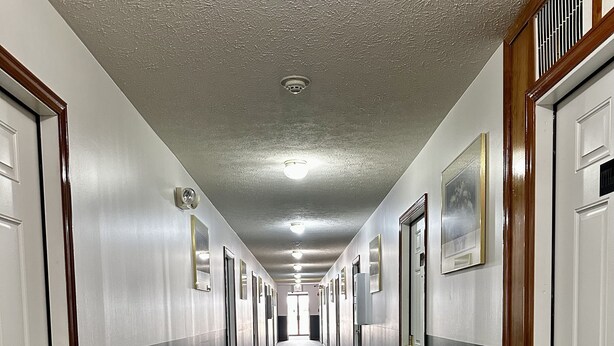 A long, narrow hallway with doors on both sides, carpeted floor, and a textured ceiling. - Attica Inn (Attica)