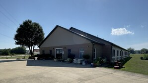 A large, single-story building with a flat roof, a paved driveway, and a lawn in front. - Attica Inn (Attica)