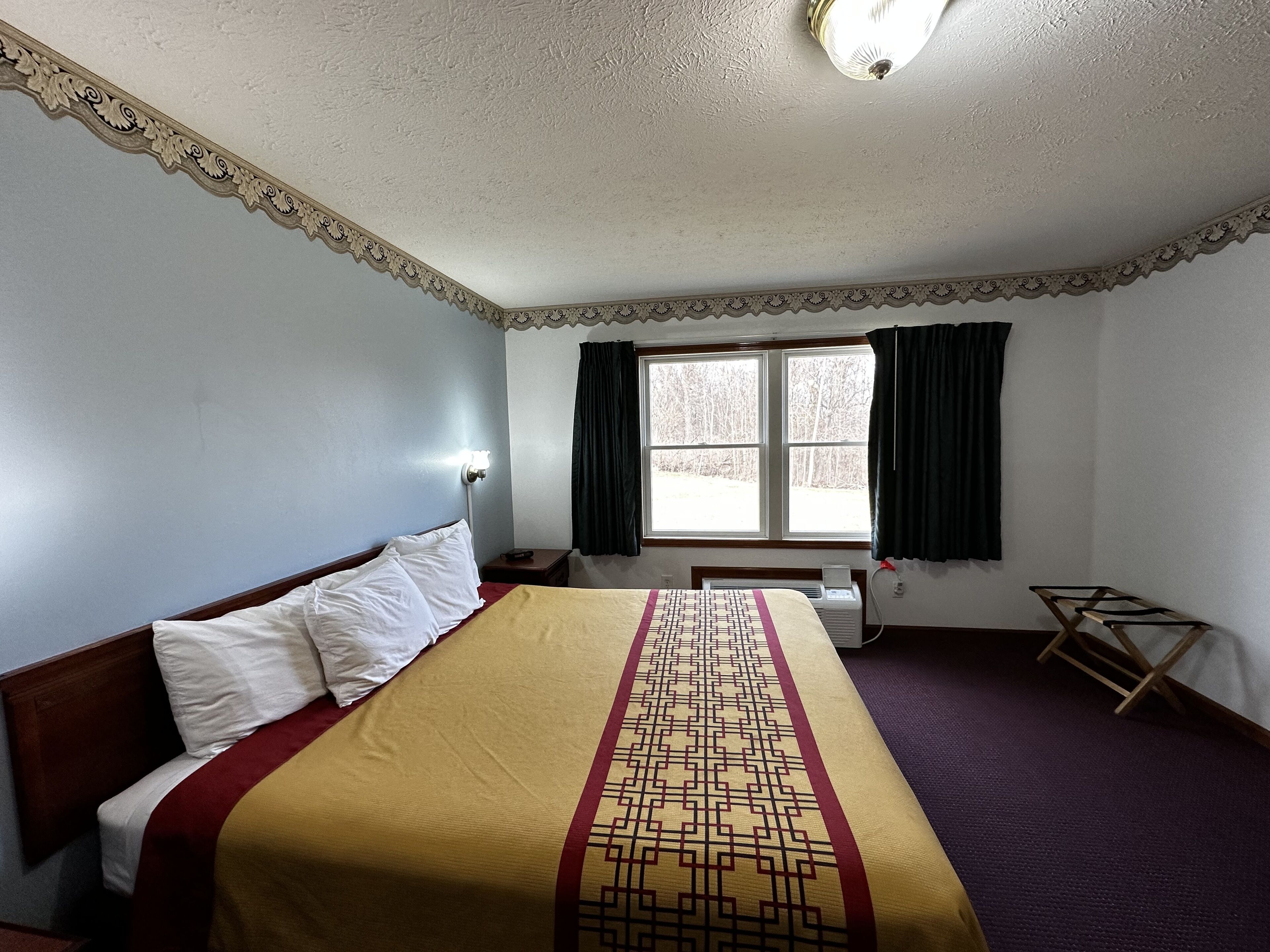 Una habitación de hotel con una cama, una ventana con cortinas, una mesita de noche y una lámpara fijada a la pared.