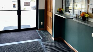 An entrance with an EXIT sign, a wooden door, a reception desk with a window, and a tiled floor with a mat. - Attica Inn (Attica)