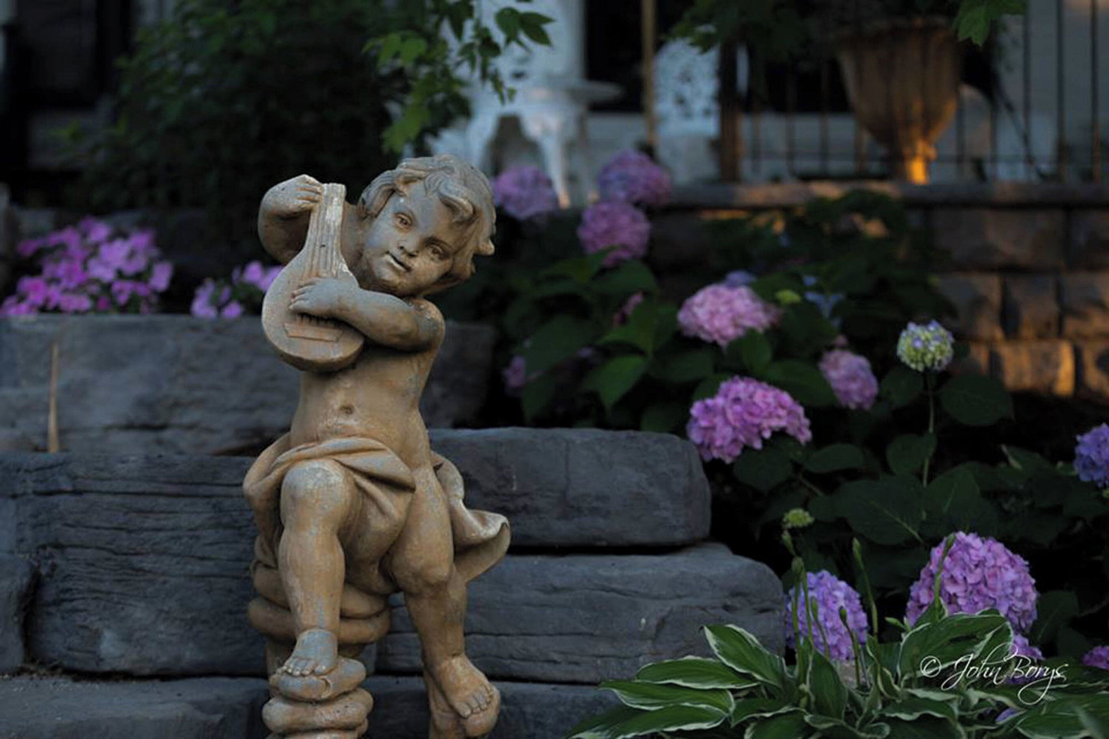 A garden statue of a child holding a lyre, surrounded by purple hydrangea flowers and a stone bench.