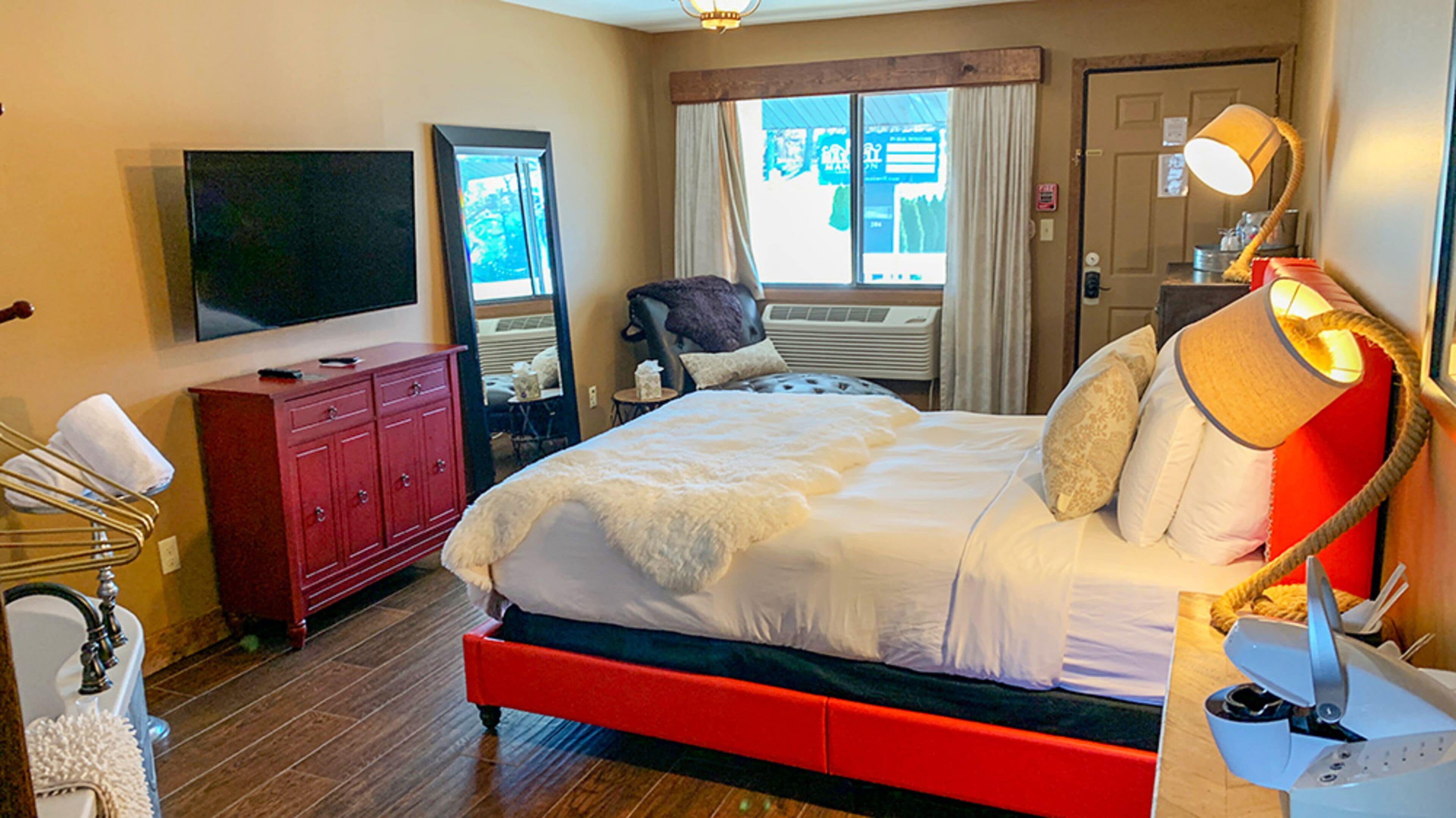 Quarto de hotel com cama grande, televisão, escrivaninha com luminária e janela com cortinas.