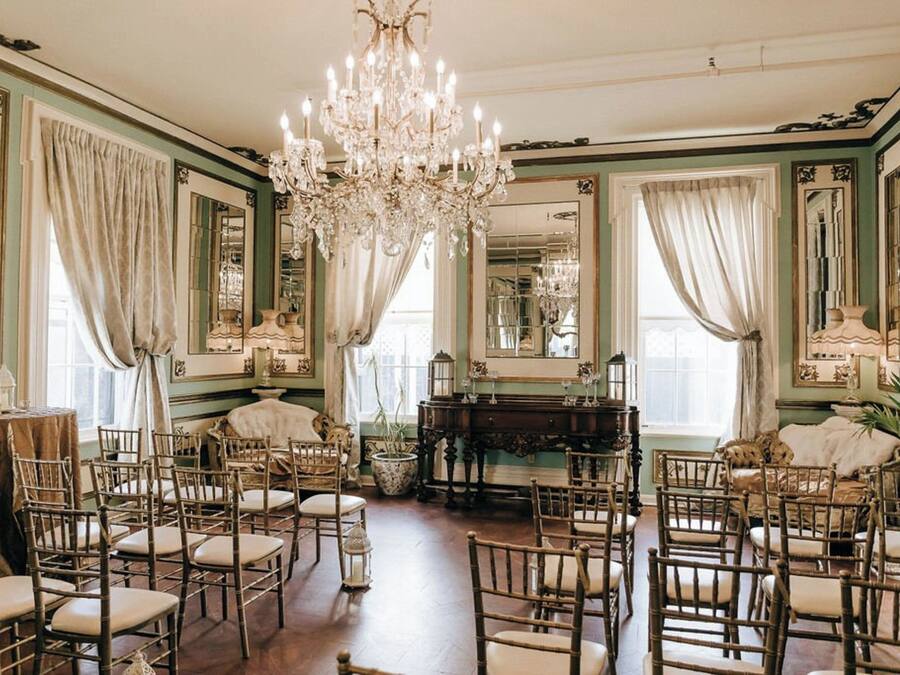 A room with a large chandelier, multiple chairs, a piano, and mirrors on the walls.