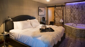 A bedroom with a large bed, a bedside table, a lamp, a tray with a mug and a glass, and a bathtub with a decorative wall. - Maxwell Mansion (Lake Geneva)