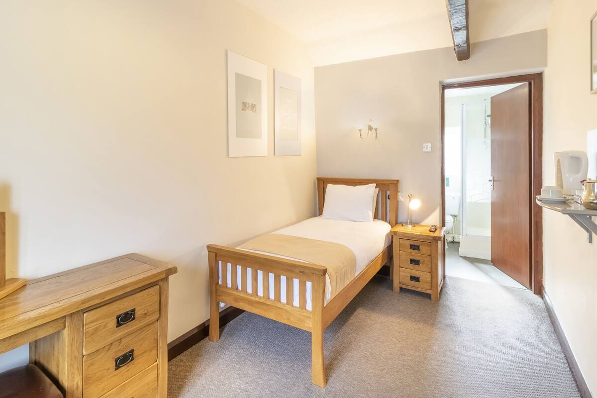A bedroom with a wooden bed, bedside tables, a desk, and a chair. There are framed pictures on the wall and a door leading to another room.