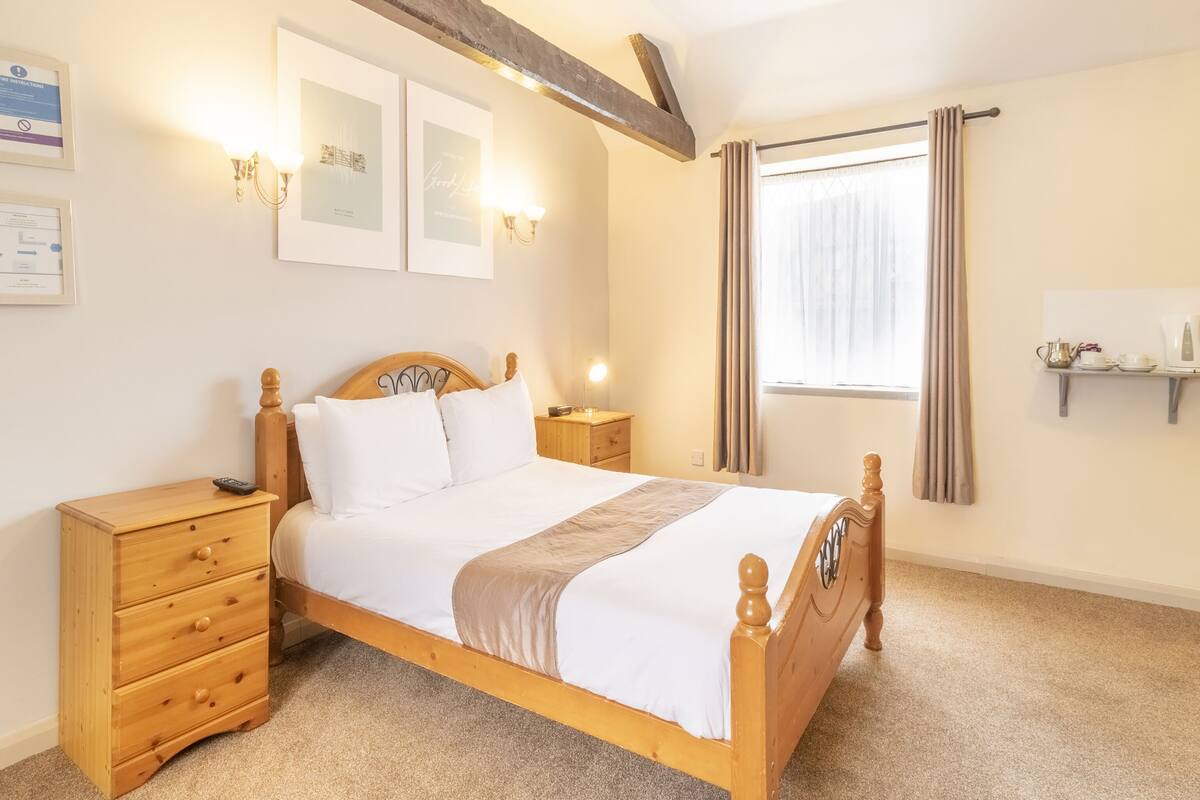 A neatly arranged bedroom with a wooden bed, bedside tables, a nightstand, a window with curtains, and wall-mounted light fixtures.