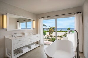 A modern bathroom with a large bathtub, a white vanity with a mirror, and a view of the beach. - Le Soleil d'Or (West End)