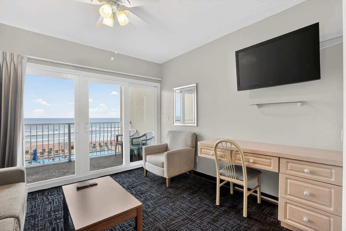 Habitación de hotel con balcón, escritorio, silla, televisor y vista a la playa.