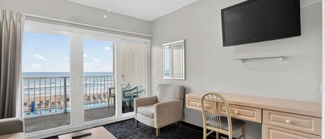 A hotel room with a balcony, a desk, a chair, a television, and a view of the beach.