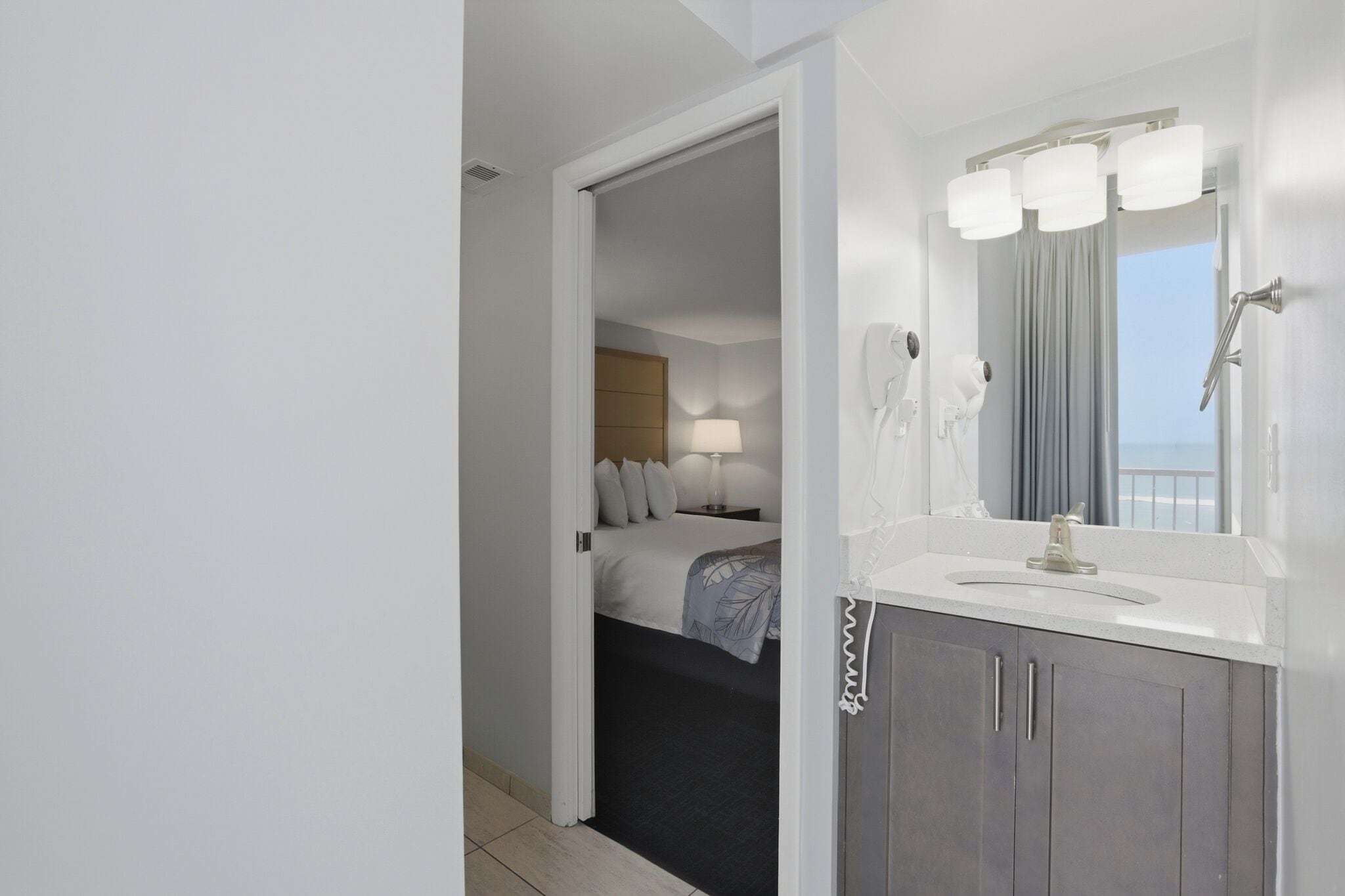 A hotel room with a bed, a bathroom with a sink and mirror, and a view of the sea.