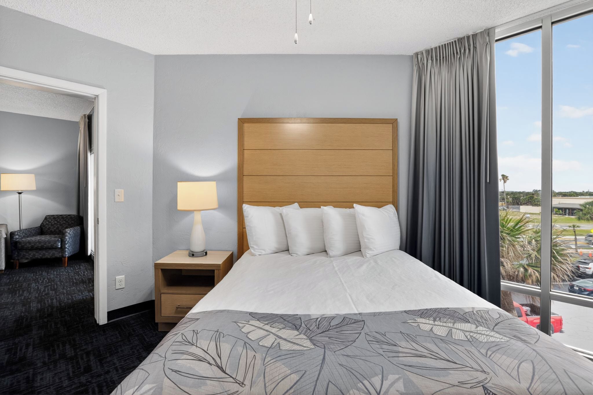 A hotel room with a bed, a nightstand, a lamp, a chair, and a view of the parking lot.