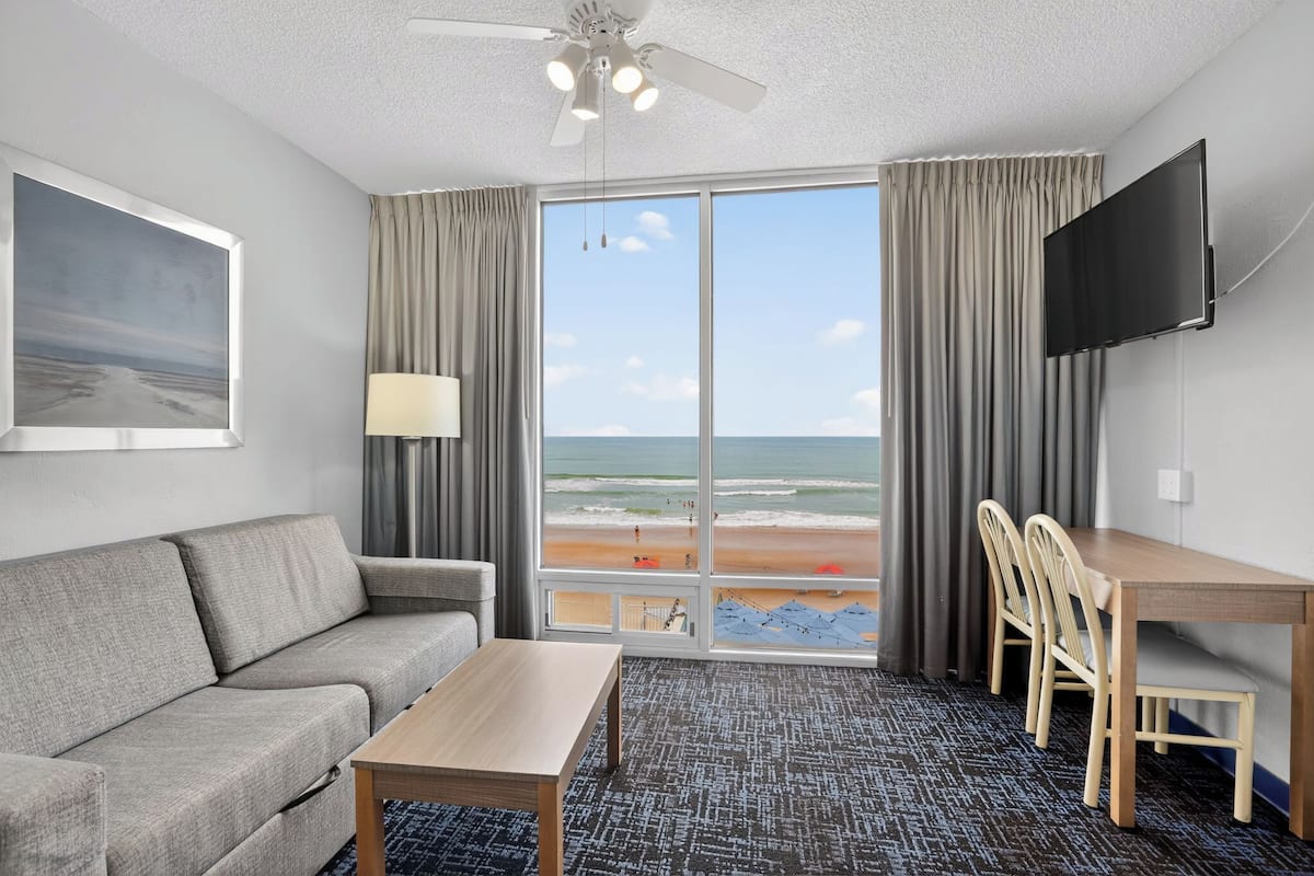 Habitación de hotel con sofá, mesa, televisión y una ventana grande con vista a la playa.