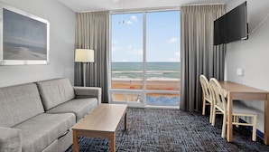 Une chambre d’hôtel avec un canapé, une table, une télévision et une grande fenêtre donnant sur la plage.