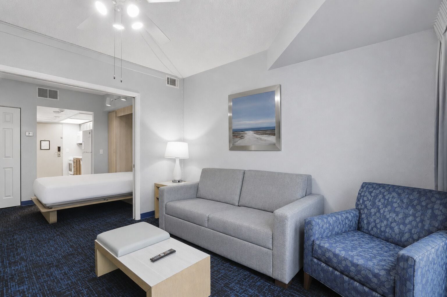 A hotel room with a bed, a grey sofa, a blue patterned armchair, a small table with a remote, a lamp, and a framed picture on the wall.