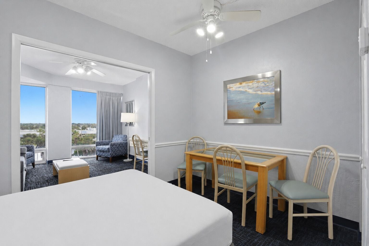 A hotel room with a bed, a dining table with chairs, a painting on the wall, and a view through a door.
