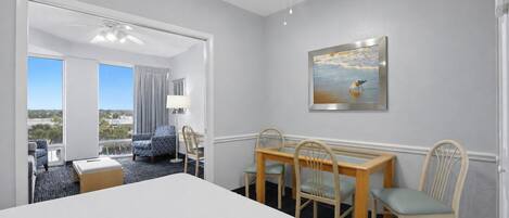 A hotel room with a bed, a dining table with chairs, a painting on the wall, and a view through a door.