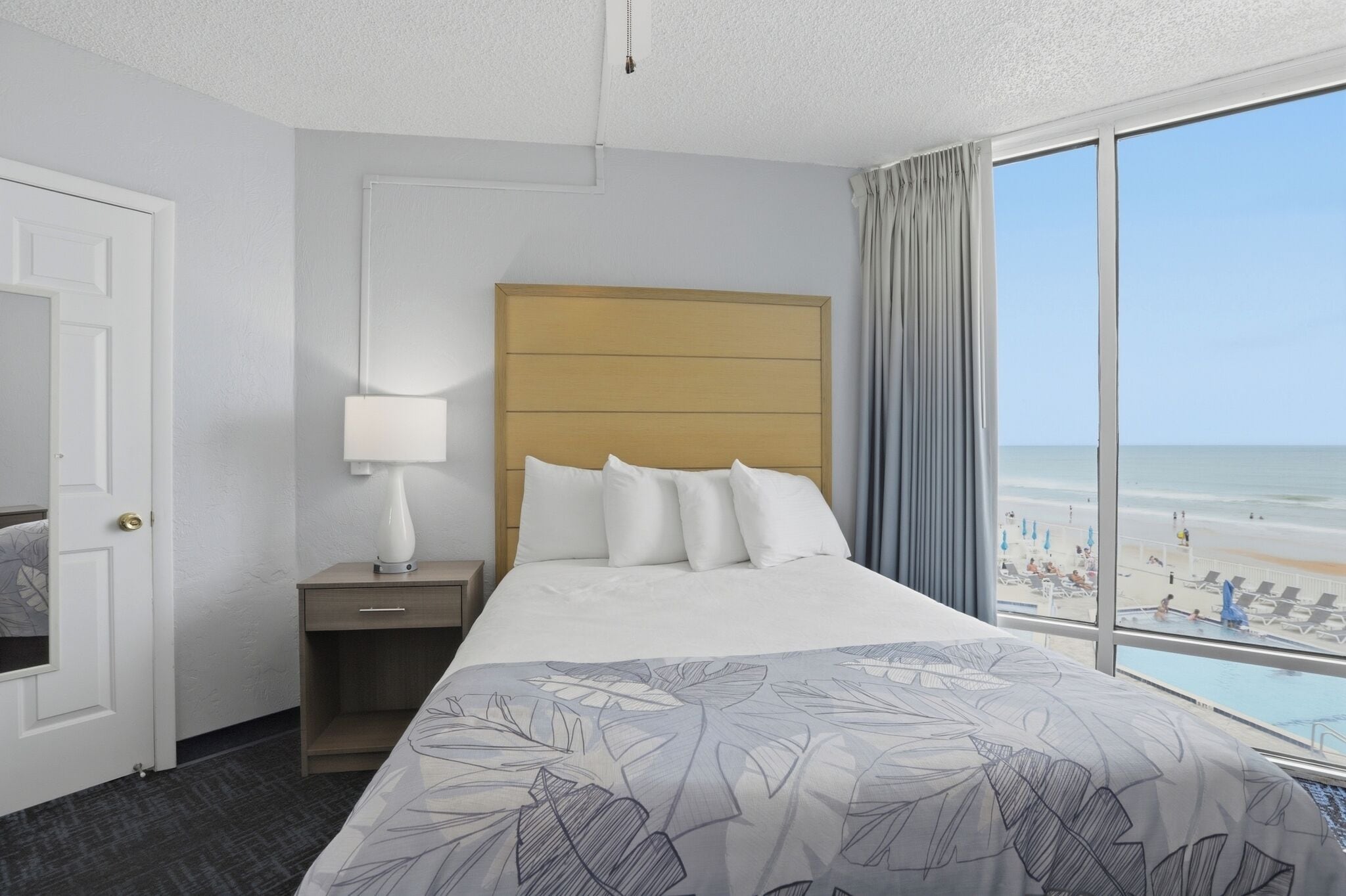Habitación de hotel con cama, mesita de noche, lámpara y vista a la playa, además de una puerta.