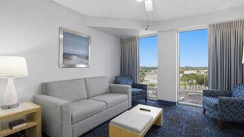 A hotel room with a grey sofa, a patterned armchair, a wooden coffee table, a lamp, and a large window with a view of the city.