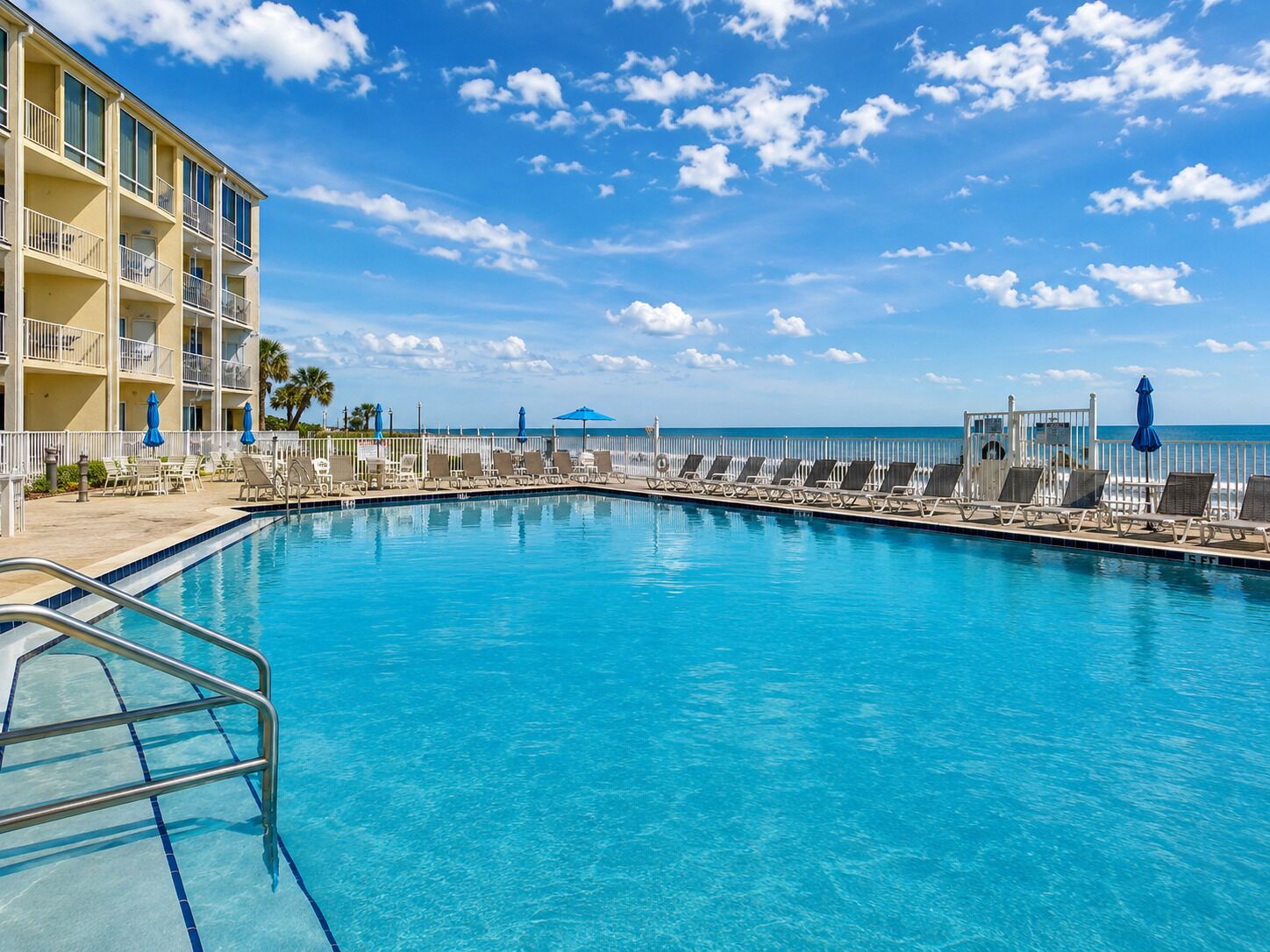 A swimming pool with lounge chairs and umbrellas, a building with balconies, and a view of the ocean.