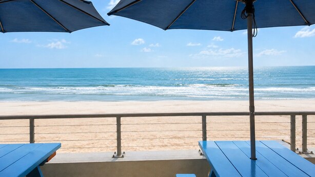 Une vue sur la plage avec deux parasols bleus et des tables de pique-nique en bois.