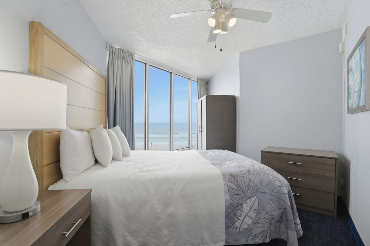 Habitación de hotel con cama, mesita de noche, cómoda, ventilador de techo y ventana grande con vista a la playa.