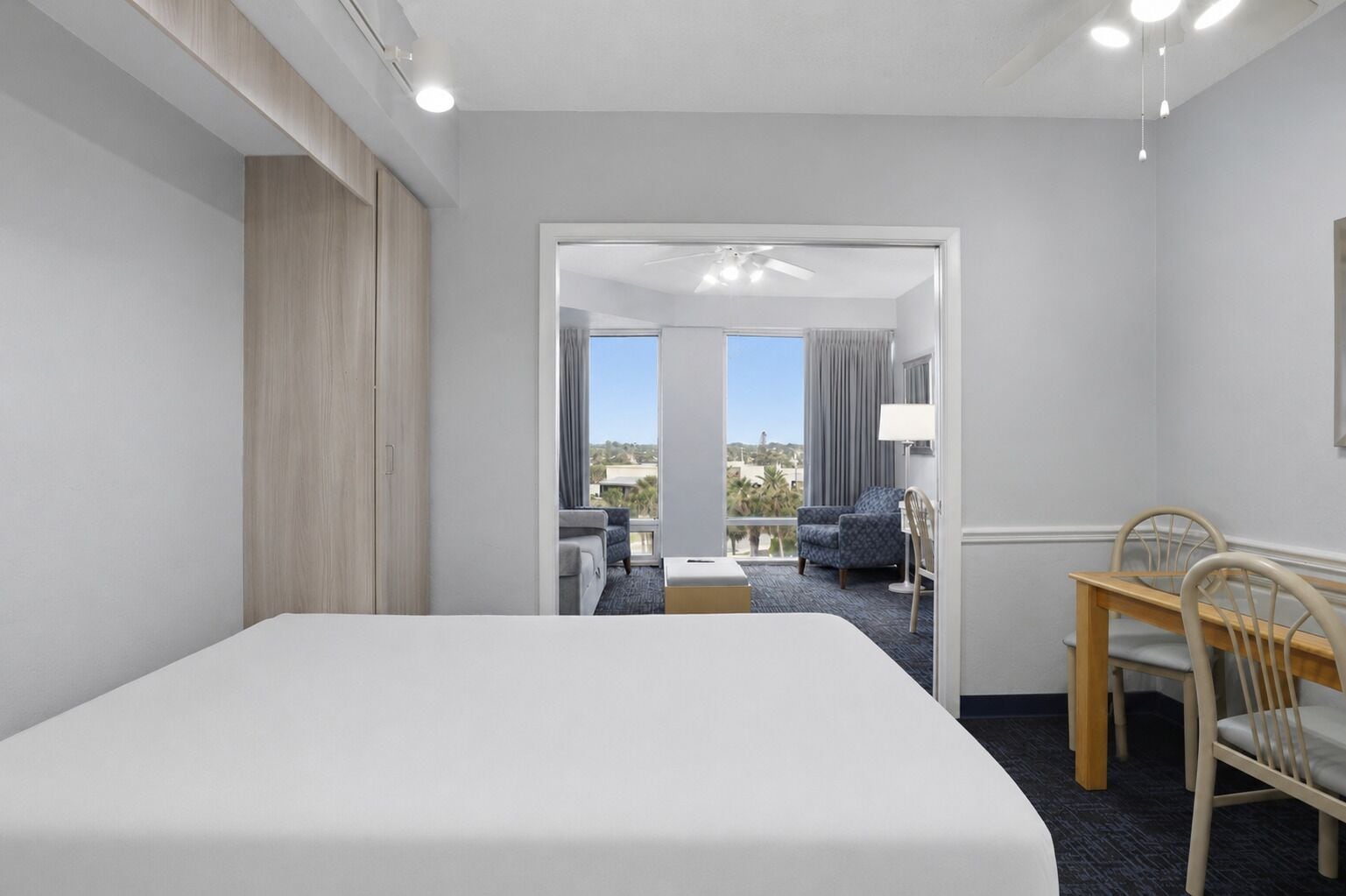 A hotel room with a large bed, a desk with chairs, a view of the outdoors through a door, and a ceiling fan.