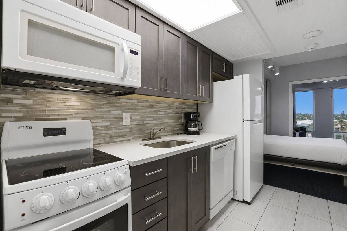 Una cocina compacta con microondas, estufa, pileta y lavavajillas, junto con un balcón pequeño con cama.