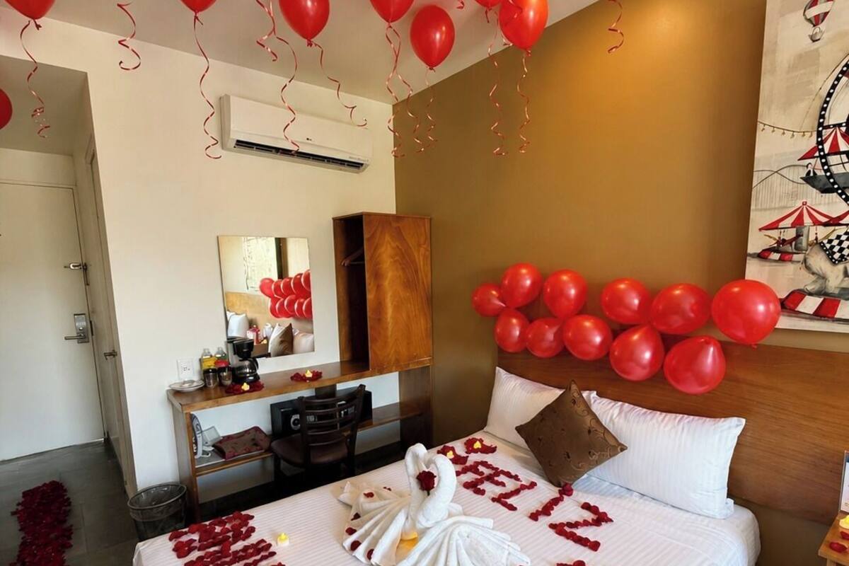A hotel room with a bed adorned with rose petals and a swan towel, red balloons, and a wall painting.