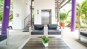 Un área de estar al aire libre techada con dos bancos acolchados, una mesita con una planta y una escalera que conduce a un ascensor.