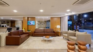 A modern hotel lobby with a brown leather sofa, a round coffee table, and a TV mounted on the wall. - Tri Hotel Executive Brusque (Brusque)