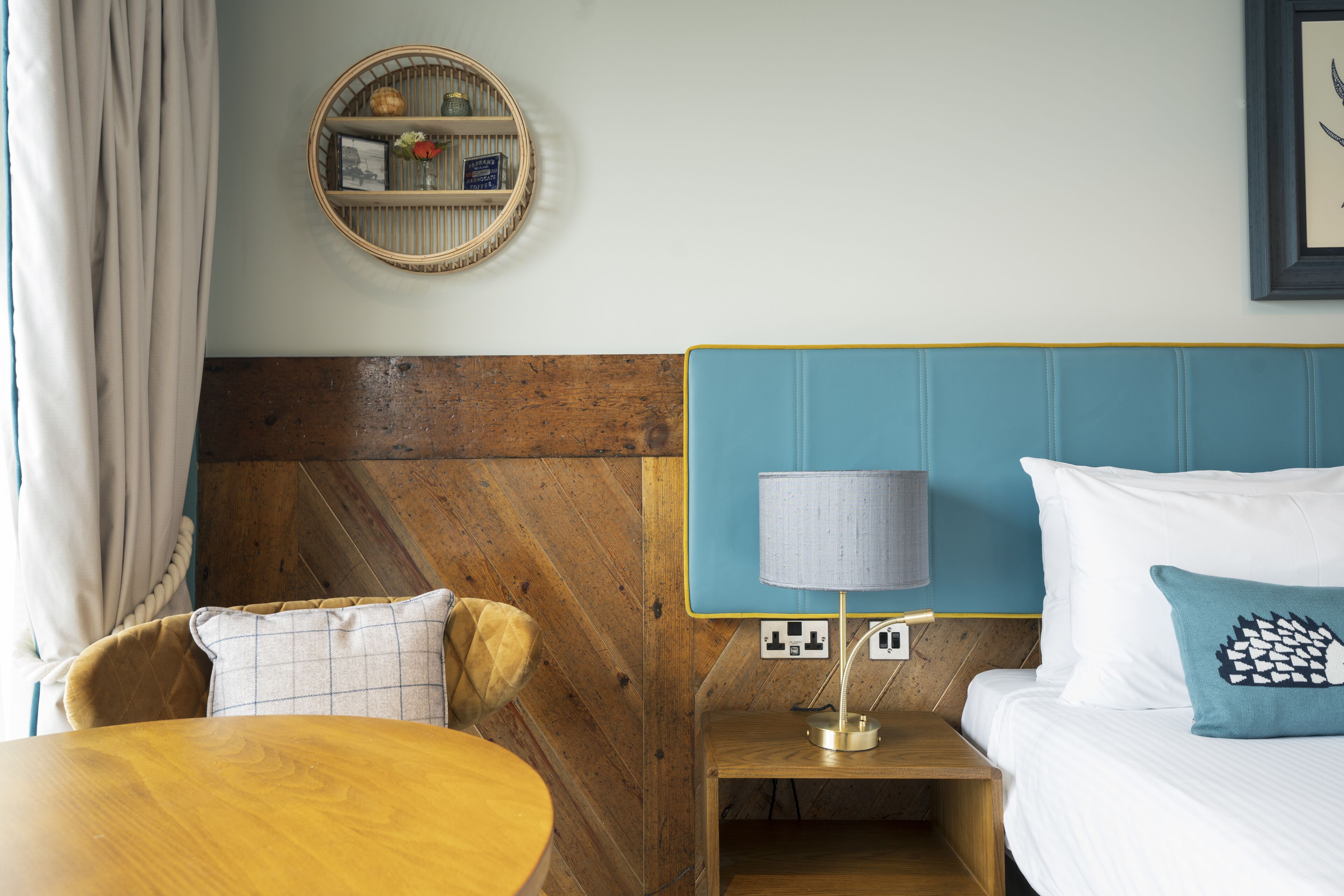 A hotel room with a wooden headboard, a round table, a bedside lamp, and a wall-mounted shelf with decorative items.