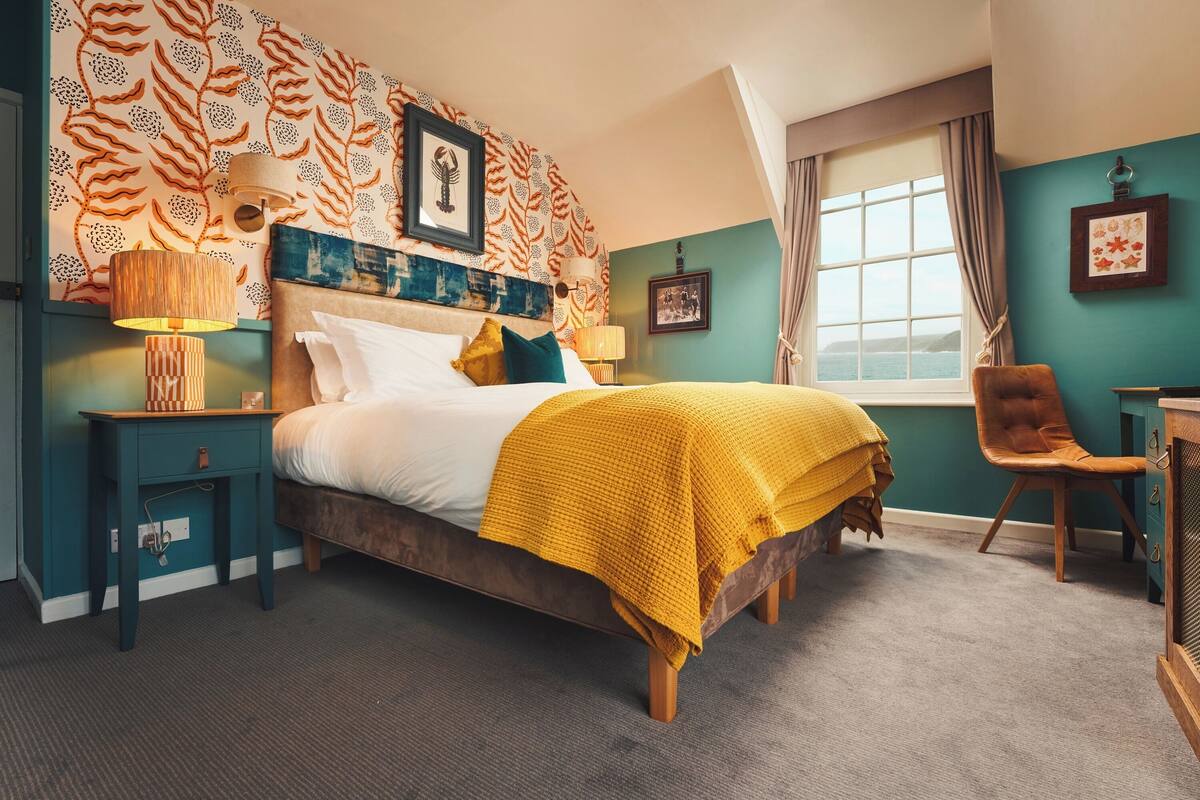 A bedroom with a bed, a nightstand, a chair, a window with a view, and a wall with patterned wallpaper.