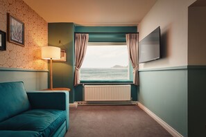 A room with a teal sofa, a window with curtains, a lamp, and a TV on the wall.