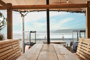 An outdoor seating area with wooden tables and chairs, overlooking a body of water and distant hills.