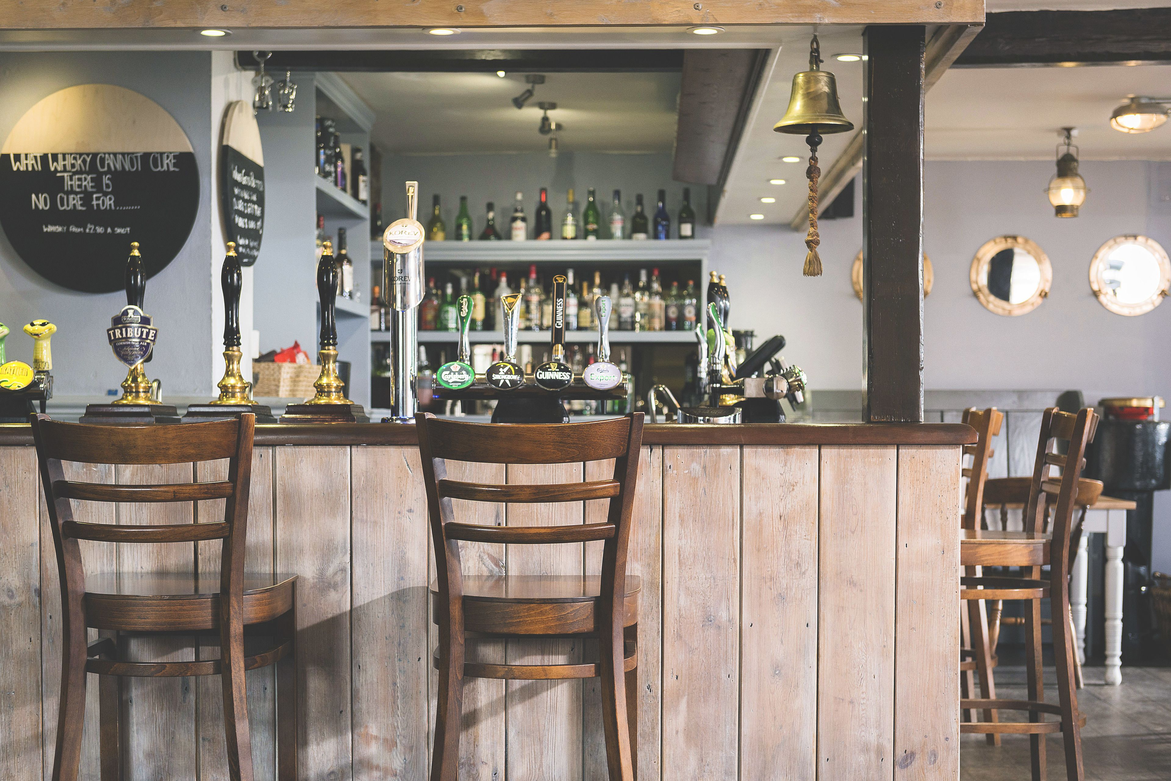 A bar area with wooden chairs, a bar counter, and various bottles and taps.