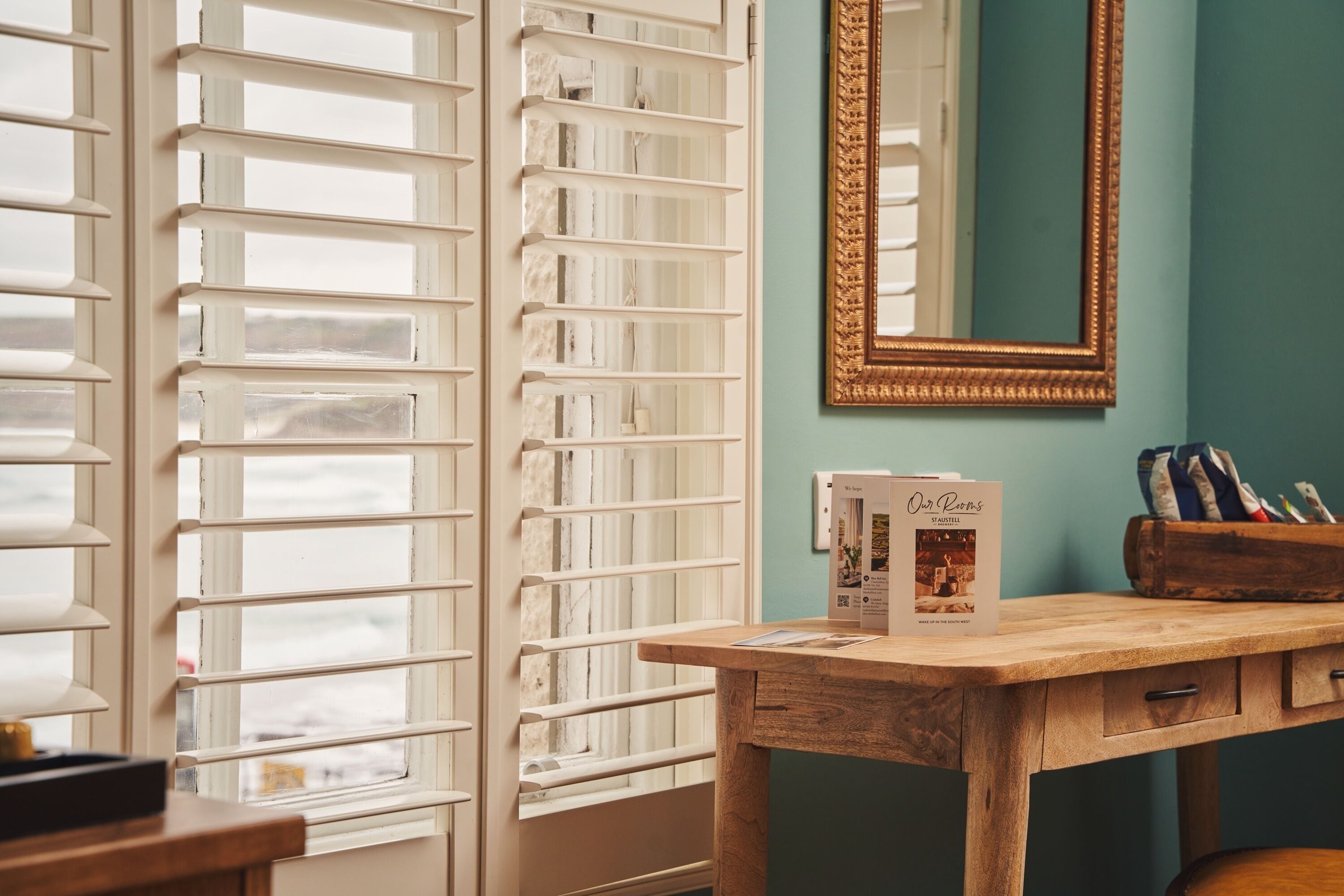 Una habitación con un escritorio de madera, un espejo con marco dorado y una ventana con persianas blancas.