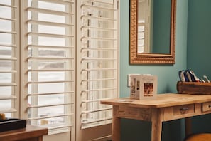 Una habitación con un escritorio de madera, un espejo con marco dorado y una ventana con persianas blancas.