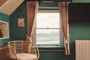 A room with a view of the sea through a window, a wicker chair, and a wall-mounted TV.