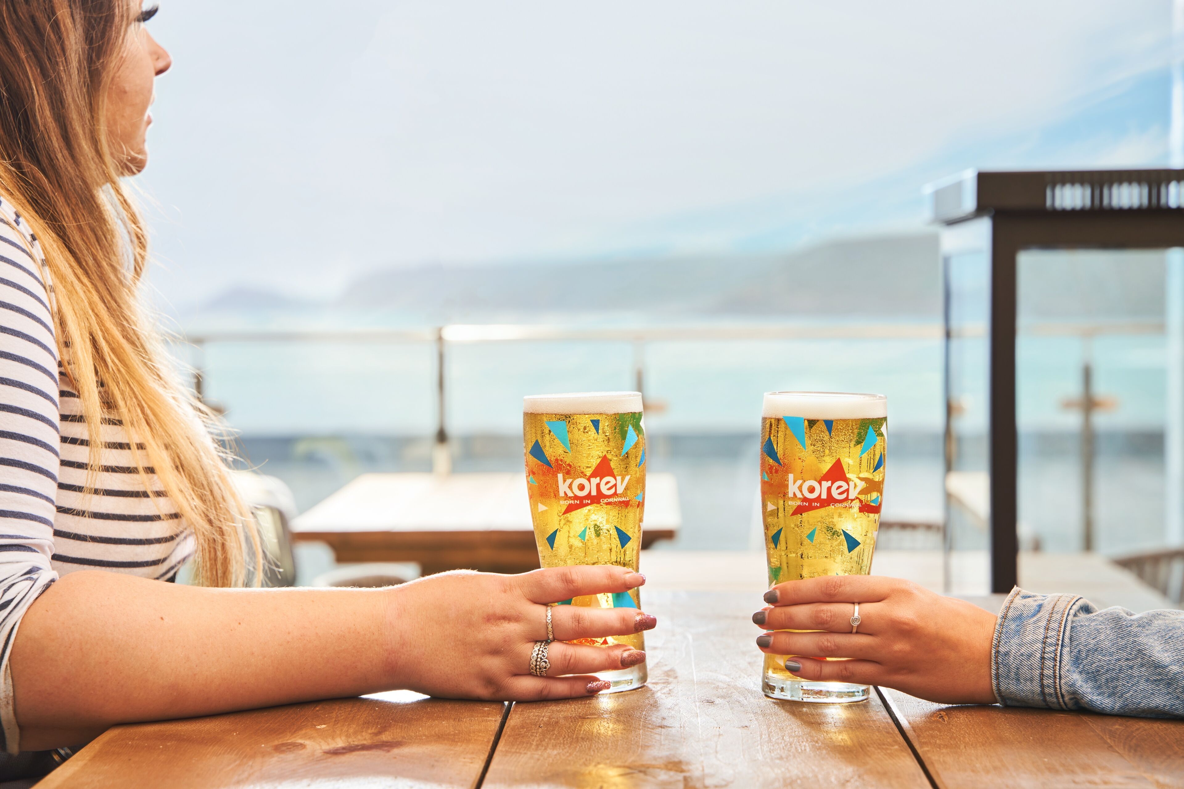 Deux personnes sont assises à une table en bois, devant deux verres de bière étiquetés « korev », dans un cadre côtier.